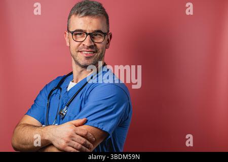 Un jeune travailleur de la santé en gommage bleu sourit avec confiance en croisant les bras. Il se tient sur un fond rose doux, mettant en valeur un et amical Banque D'Images
