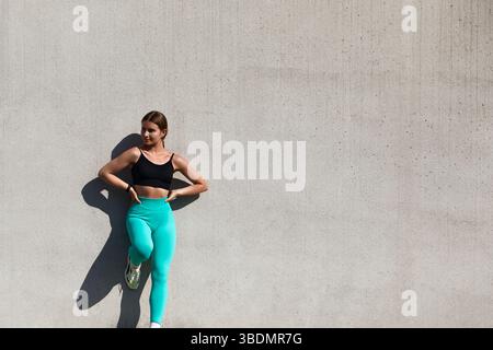 Jeune femme dans des vêtements de sport élégants se tient en toute confiance contre un mur texturé, incarnant la forme physique, le style et un style de vie actif qui inspire w Banque D'Images