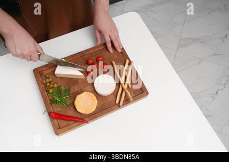 Un homme dans la cuisine coupe du fromage sur une planche sur une table blanche. Différents types de fromage, tomates, Chili, olive, roquette, couteau, bâtonnets de pain. Appetiz Banque D'Images