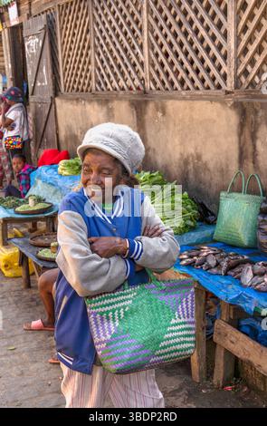 Femme malgache âgée sur un marché local animé à Madagascar, portant une veste bleue et un sac tissé vert, souriant comme elle se tient devant des produits frais an Banque D'Images