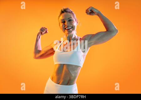 Belle femme sportive mature portant des vêtements de sport faisant de l'entraînement - athlète senior heureux fléchissant biceps en vêtements de sport, souriant contre un dos orange Banque D'Images