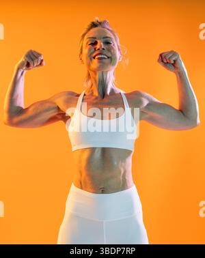Belle femme sportive mature portant des vêtements de sport faisant de l'entraînement - femme mature sportive fléchissant les muscles en tenue athlétique sur un fond vibrant Banque D'Images