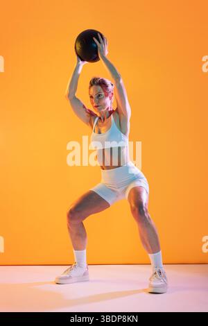 Belle femme sportive mature portant des vêtements de sport faisant de l'entraînement - femme mature énergique dans des vêtements de sport soulevant un medicine ball au-dessus de la tête tout en squatti Banque D'Images