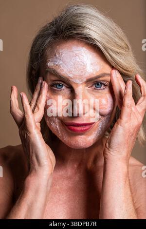 Portrait de beauté de belle femme mature, concepts sur les cosmétiques, les soins de la peau et le bien-être. Heureuse femme mature appliquant la crème hydratante sur son visage Banque D'Images