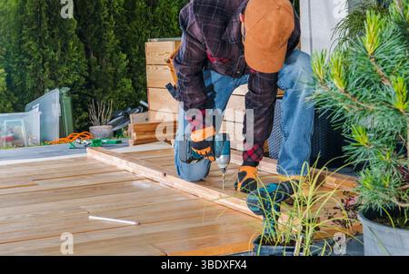 Une personne s'agenouille sur une terrasse en bois partiellement finie, en utilisant une perceuse électrique pour fixer les planches. Une végétation luxuriante entoure la zone de travail, indiquant Banque D'Images