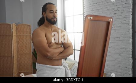 Homme musclé avec barbe se tient en toute confiance avec les bras croisés, enveloppé dans une serviette, reflétant dans le miroir pleine longueur dans l'intérieur moderne de la maison avec naturel Banque D'Images