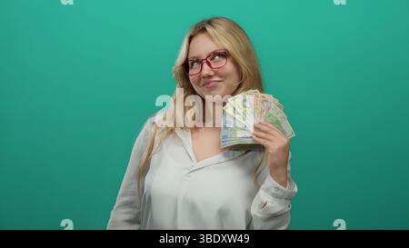 Jeune femme dans des lunettes tenant des billets de banque roumains lei contre un mur vert, exsudant confiance et richesse, isolée sur un fond vivant pour financia Banque D'Images