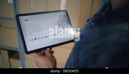 Employé vérifiant les actions en temps réel et les graphiques du marché des changes à l'aide de Digital Tablet Computer. Homme faisant des investissements pendant les heures de travail dans l'entrepôt, combinant l'emploi avec le commerce en ligne. Gros plan. Banque D'Images
