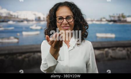 Femme avec les cheveux bouclés étend la main à la caméra à l'extérieur dans un port de bord de mer avec des bateaux flottant en arrière-plan sur une journée ensoleillée. Banque D'Images