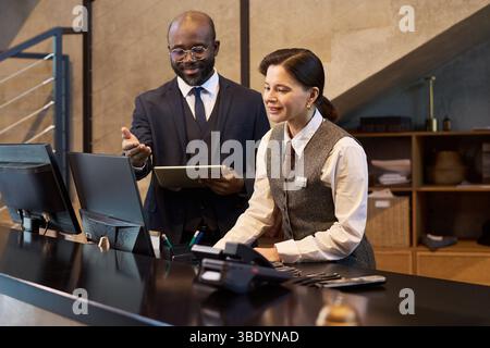 Deux professionnels debout à la réception discutant des détails du travail à l'aide de la technologie et de la documentation. Homme tenant la tablette tandis que femme prenant des notes au bureau Banque D'Images