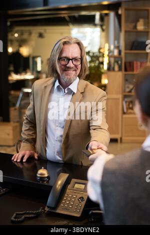 Portrait d'un vieil homme souriant avec des lunettes tenant une carte de crédit à la réception de l'hôtel debout en face du personnel. Arrière-plan comprend des décorations et de l'équipement de bureau Banque D'Images