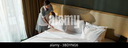Femme de ménage ajustant les oreillers et faisant le lit dans une chambre d'hôtel moderne. Les draps blancs soigneusement disposés créent une atmosphère propre et accueillante Banque D'Images