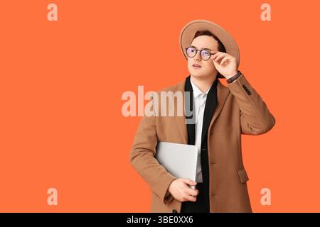 Jeune homme en chapeau, manteau et avec ordinateur portable sur fond orange Banque D'Images