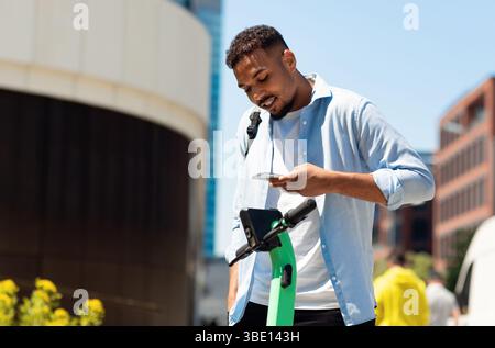 Location d'un scooter électrique. Jeune afro-américain louant un transport écologique, payant en ligne avec un téléphone portable Banque D'Images