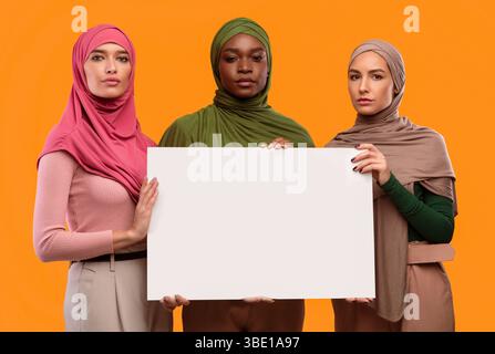Trois femmes musulmanes graves tenant un tableau de papier vierge, fond jaune Banque D'Images