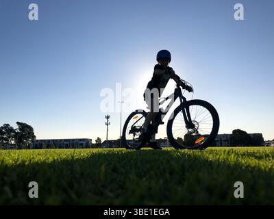 Silhouette d'enfant debout au-dessus de leur VTT sur le terrain d'herbe au coucher du soleil, appréciant le style de vie en plein air d'été Banque D'Images