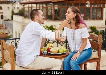 couple est assis à une petite table en bois sur un toit-terrasse, partageant un petit déjeuner de fruits frais. Ils semblent détendus et ludiques, appréciant les uns les autres comp Banque D'Images