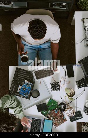 Deux individus assis en face l'un de l'autre à l'établi entouré de composants électroniques démontés et de pièces d'ordinateur, l'un tenant un café, d Banque D'Images