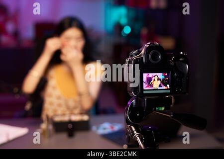 Femme filmant tutoriel de maquillage tout en étant filmé par une caméra professionnelle en studio, avec fond néon violet flou. Influenceuse féminine créant du contenu pour vlog Banque D'Images