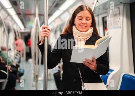 Une femme enthousiaste englouti dans un livre intrigant tout en se tenant debout dans un train de métro, pleinement absorbée par son expérience de lecture et le passage du temps Banque D'Images