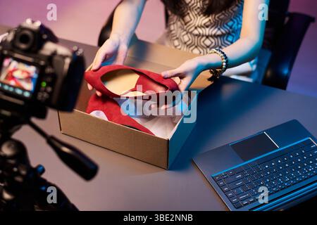 Femme anonyme assise au bureau déballant et examinant les chaussures rouges à talons hauts à la mode, avec caméra sur le processus d'enregistrement trépied, avec ordinateur portable à proximité Banque D'Images