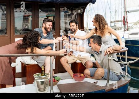 Six amis adultes célébrant sur le pont d'un bateau à moteur, toast avec du vin mousseux et riant lors d'une croisière estivale dans la marina. Concept de Banque D'Images