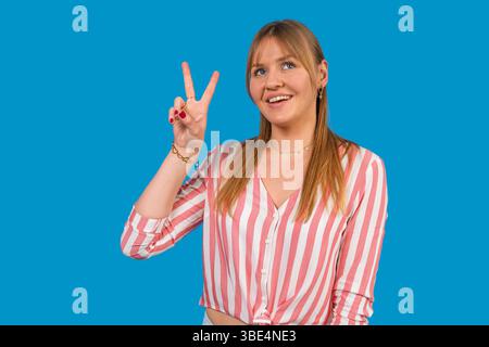 Jeune femme joyeuse gestant la victoire avec deux doigts, souriant et regardant loin sur un fond bleu Banque D'Images
