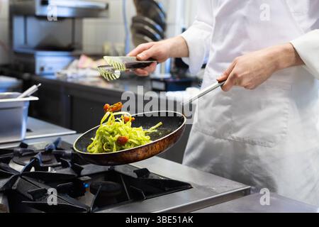 Deutschland 07. Avril 2025 : Im Bild : Ein Koch in Weißer Arbeitskleidung schwenkt gekonnt Pasta mit getrockneten Tomaten und grüner Soße in einer rustikalen Pfanne über einem Gasherd in einer professionellen Restaurantküche. Die Bewegung des Schwenkens ist dynamisch eingefangen, wobei die Nudeln in der Luft sind und das können des Kochs unterstreichen. Der Hintergrund zeigt eine typische, moderne Küchenumgebung mit Edelstahloberflächen und professionellen Geräten. Dieses Bild eignet sich Ideal für Kochen, gastronomie, Restaurantbetrieb, Küchenpersonal, Kochkünste, Zubereitung von Speisen, itali Banque D'Images