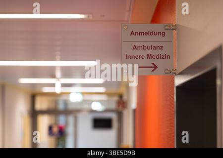 Deutschland 25. Avril 2025 : Im Bild : Ein gut sichtbarer Wegweiser aus Metall oder Plexiglas ist in einem modernen Krankenhausflur angebracht und zeigt die Richtungen zu Anmeldung , Ambulanz und Innere Medizin mit einem deutlichen Pfeil nach rechts. Der Flur ist Hell beleuchtet mit Reihen von Deckenleuchten und die Wände sind in neutralen Farbtönen gehalten, teilweise mit einem Akzent in Orange. Die Szene vermittelt den Eindruck eines organisierten und leicht zugänglichen Bereichs innerhalb einer medizinischen Einrichtung. Dieses Foto eignet sich hervorragend für Verwendungen zum Thema Gesundhe Banque D'Images