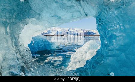 Vue imprenable de l'intérieur d'une grotte de glace bleu cristal à Jokulsarlon, Islande, encadrant le lagon glaciaire et les icebergs flottants avec des montagnes enneigées Banque D'Images