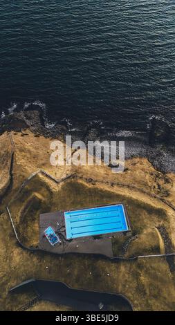 Photo aérienne unique d'une piscine bleu vif située sur la côte islandaise accidentée, contrastant avec les eaux sombres de la mer et de la terre Banque D'Images