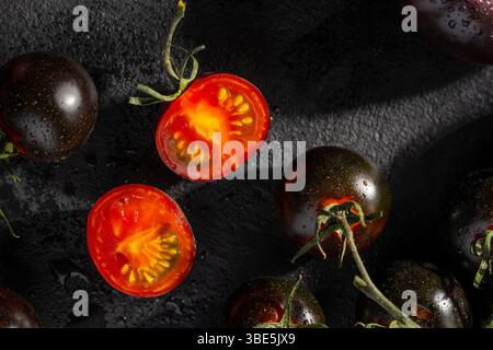 Un groupe de tomates cerises noires entières et coupées en deux, présentées sur une surface texturée sombre, mettant en valeur leur peau brillante et leur intérieur rouge vif. Banque D'Images