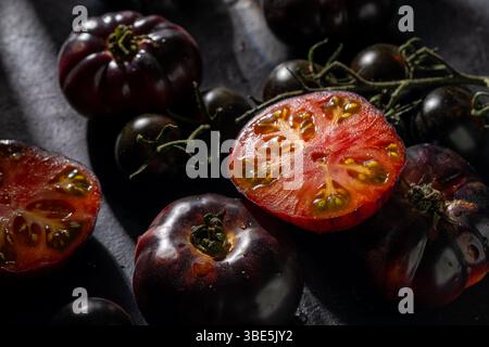 Les tomates noires, entières et tranchées, affichent des intérieurs rouges vifs sur une surface sombre, éclairées par un éclairage spectaculaire. Un goût d'élégance et naturel f Banque D'Images