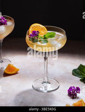 Cocktails dans deux verres est élégamment garni d'une tranche d'orange, de fleurs violettes vibrantes et de feuilles de menthe fraîche, créant un équilibre parfait de col Banque D'Images
