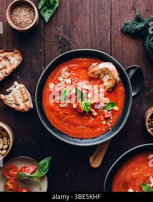 Un bol de soupe vibrante aux tomates garni de feuilles de basilic frais et de graines de sésame est servi sur une table en bois rustique, accompagné d'un morceau de pain croustillant Banque D'Images