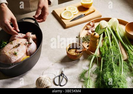 Un cuisinier assaisonne un poulet entier avec des herbes fraîches et du citron dans un cadre de cuisine. Le fenouil, l'ail et d'autres assaisonnements sont préparés sur une table pour une fla Banque D'Images