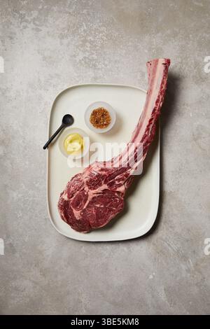 Vue de haut en bas d’un steak de tomahawk cru présenté sur un plateau en céramique, accompagné d’un petit bol de moutarde, d’épices et d’une cuillère noire. Une coupe premium réf Banque D'Images