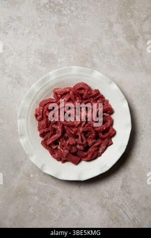 Une vue de haut en bas d'une assiette en céramique remplie de lamelles de bœuf fraîchement coupées, soigneusement disposées sur une surface texturée neutre, idéale pour cuisiner de l'adepsie Banque D'Images