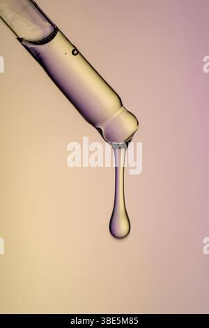 Plan macro d'un compte-gouttes de sérum suspendu à une seule goutte, capturé sur un fond dégradé rose et jaune doux. Banque D'Images