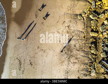 Vue aérienne d'une plage de sable avec de longues ombres projetées par des personnes et des chiens marchant en Australie. Des empreintes de pas traînent derrière eux, et des formations rocheuses alignent t Banque D'Images
