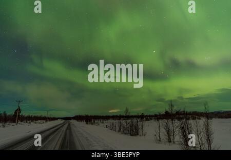 Vue imprenable sur les aurores boréales illuminant le ciel nocturne avec des teintes vertes vibrantes au-dessus d'une route enneigée et d'un paysage près de Kiruna, en Suède. Banque D'Images
