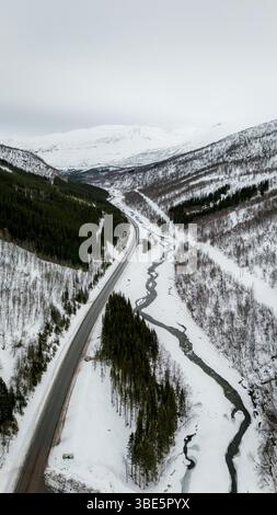 Vue aérienne d'un paysage nordique avec une route traversant des montagnes et des forêts enneigées, mettant en valeur la beauté sereine d'un pays des merveilles hivernales Banque D'Images