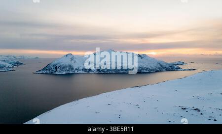 Une vue aérienne à couper le souffle capture un paysage hivernal nordique avec une île enneigée tranquille entourée d'eaux calmes sous un ciel doux et pastel coucher de soleil, Banque D'Images