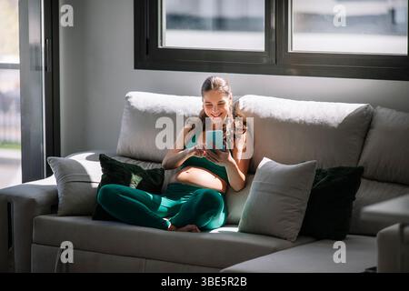 Une femme enceinte est assise confortablement sur un canapé confortable, portant une tenue de yoga. Elle aime son temps libre, se concentrant sur le bien-être et se connectant numériquement th Banque D'Images