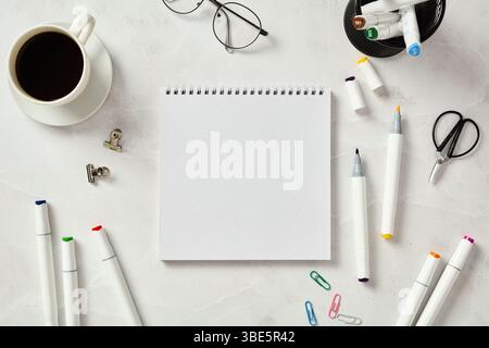 Vue de dessus d'un carnet de croquis blanc entouré de marqueurs colorés, une tasse de café, des verres et des fournitures de bureau, soigneusement disposés sur une table en marbre. Banque D'Images