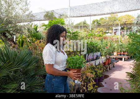 Heureuse femme afro-américaine souriante avec des cheveux bouclés, aime sélectionner des plantes dans un centre de jardinage. Vêtue d'un élégant haut blanc et d'un Jean bleu, elle e Banque D'Images