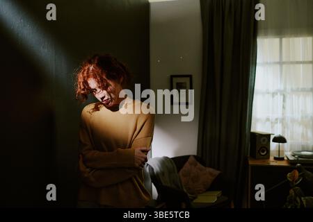 Femme aux cheveux roux profitant d'un moment calme à l'intérieur, se serrant dans ses bras. Cette chambre bénéficie d'un éclairage doux, créant une atmosphère sereine Banque D'Images
