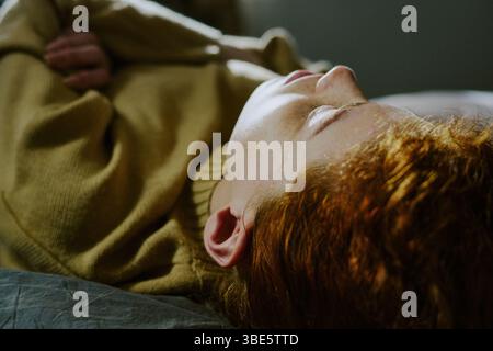 Personne allongée sur le côté sur le lit, montrant une déchirure visible sur le visage et portant un pull brun. Moment émotionnel capturé dans un intérieur confortable avec Lig doux Banque D'Images