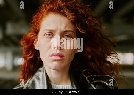 Portrait d'une femme aux cheveux rouges sous le pont portant une veste en cuir et regardant directement la caméra avec une expression sérieuse Banque D'Images