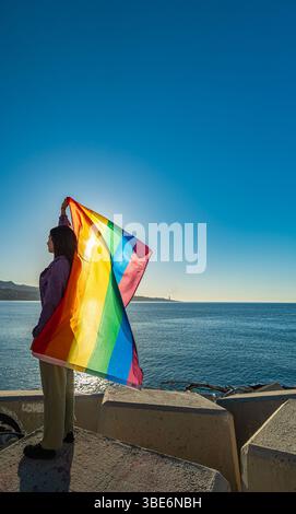 Plan lumineux d'une fille de profil levant fièrement un drapeau arc-en-ciel alors que le vent le déploie. Diversité, visibilité LGTBI et autonomisation des jeunes dans un natur Banque D'Images